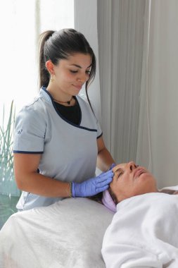 Üniformalı genç bir estetisyen, temiz bir spa ortamında olgun bir bayan müşteriye profesyonel yüz bakımı yapıyor..