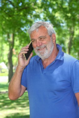 Üst düzey Latin bir adam yeşil bir parkta cep telefonuyla konuşuyor..