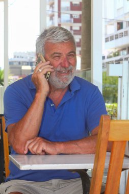 Üst düzey Latin bir adam açık bir kafede oturuyor, rahat bir ortamda kahve ya da çay içiyor, cep telefonu kullanıyor..