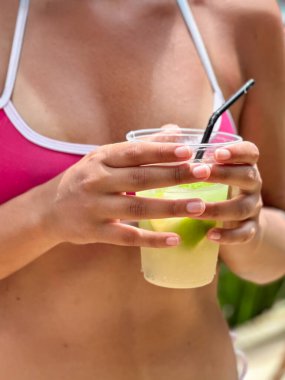 Kireç ve buzla taze bir Caipirinha kokteyli tutan kadın ellerinin yakın çekimi, tropikal bir sahil bölgesinde çekilmiş..