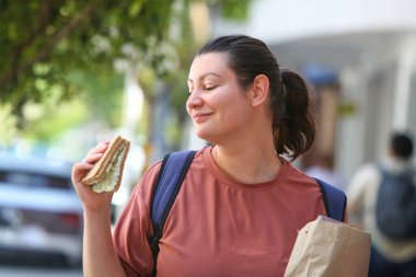 Şehrin caddesinde yürürken paket sandviç yiyen yetişkin bir kadının samimi şehir tarzı portresi..
