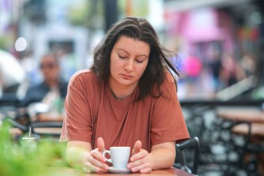 Düşünceli yetişkin kadın şehir caddesinde açık bir kafede tek başına oturuyor, önünde bir fincan kahveyle. .