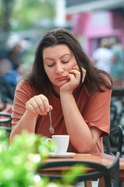 Düşünceli yetişkin kadın şehir caddesinde açık bir kafede tek başına oturuyor, önünde bir fincan kahveyle. .