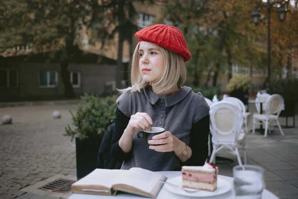 Young woman with a beret reading a book in a cafe while drinking coffee and eating cake. French street style.