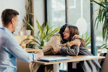 Neşeli üniversite arkadaşları kampüs kafeteryasında buluşuyor, ikisi kucaklaşırken üçüncü arkadaş gülümsüyor. Dersler arasındaki yemek salonunda mutlu sosyal buluşmalar..