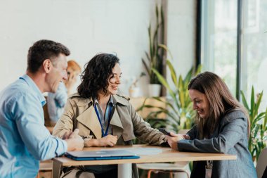 İş arkadaşları keyifli bir mola veriyor, gülüyor ve bir kafe masasında telefona bakıyorlar.