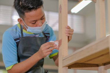 Young carpenter in work clothes and face mask  using wood filler in finishing work  for console table . clipart