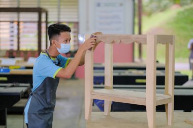 İş kıyafetleri ve yüz maskesi takan genç marangoz konsol masasının işini bitirmek için zımpara kağıdı kullanıyor. .