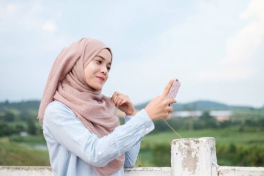 Tesettürlü neşeli ve çekici genç bir kadın selfie çekiyor. Malay modeli arka plan olarak beyaz çitlerin yanında duruyor.