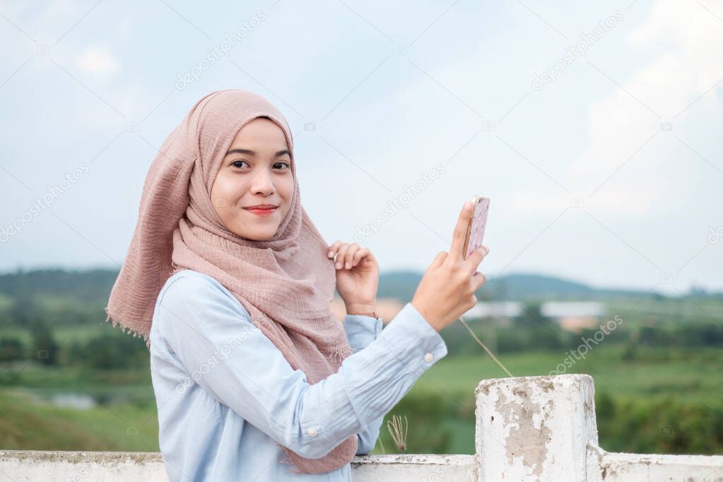 Cheerful and attractive young woman in hijab taking selfie. Malay model stands near the white fence in the farm as background