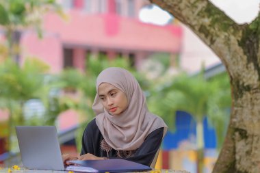 Parkta oturan ve gümüş dizüstü bilgisayar kullanan, başörtüsü takan güzel İslami kız öğrencinin fotoğrafı..