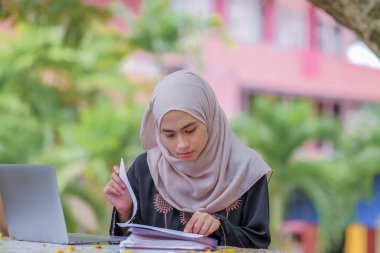 Parkta oturan ve gümüş dizüstü bilgisayar kullanan, başörtüsü takan güzel İslami kız öğrencinin fotoğrafı..