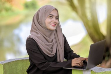 Yeşil parkta oturan ve gümüş dizüstü bilgisayar kullanan 20 'li yaşlarda güzel bir İslami öğrenci fotoğrafı..