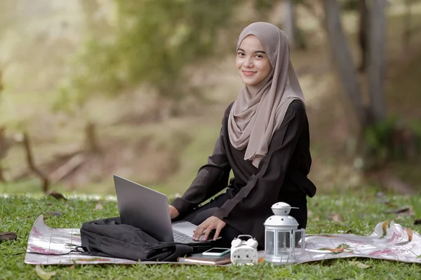 照片上美丽的伊斯兰女生20多岁 头戴头巾 坐在绿地公园里 用银色笔记本电脑 — 图库照片