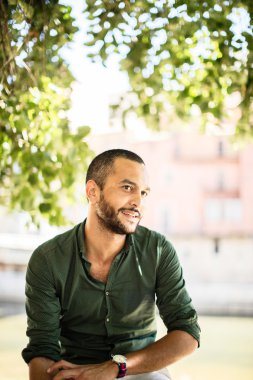 Açık havada ağaçların altında oturan ve gülümseyen bir adam genç sakallı