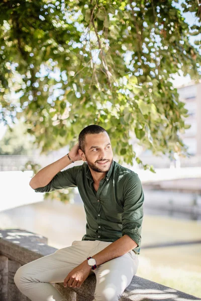 Young bearded man sitting outdoors under trees and smiling — Stock Photo, Image