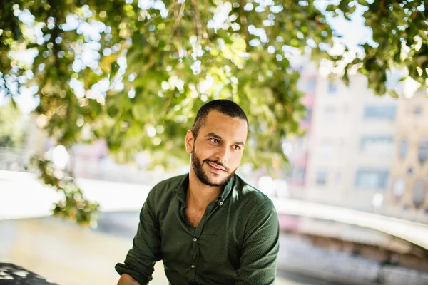 Young bearded man sitting outdoors under trees and smiling — Stock Photo, Image