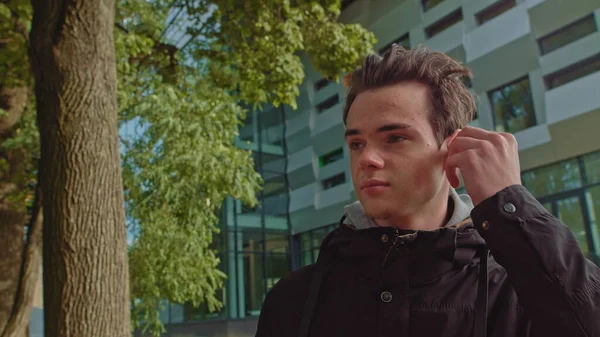 Junger Mann telefoniert mit drahtlosen Kopfhörern im Freien, während er die Straße hinuntergeht. junger Kerl, Teenager trägt moderne drahtlose Kopfhörer. Porträt eines jungen Mannes. — Stockfoto