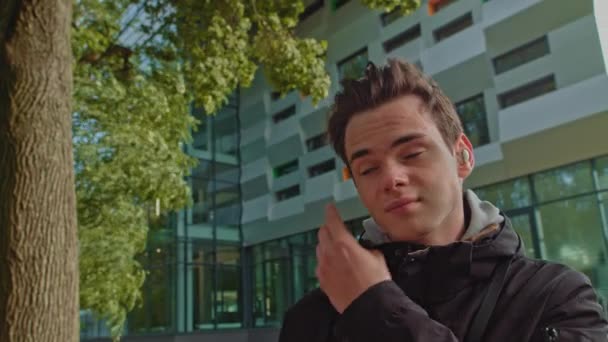 Young man talking on the phone using wireless headphones outdoors while walking down the street. young guy, teenager wears modern wireless headphones. Portrait of a young man. — Stock Video