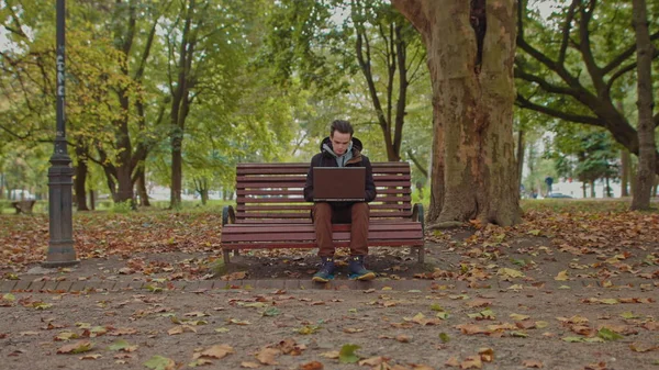 Közepes felvétel egy jóképű fiatalemberről, aki laptopon dolgozik, és a szabadban ül a parkban. A férfi figyelmesen néz félre és mosolyog. Otthoni életmódból dolgozom. karantén a Covid-19-ben — Stock Fotó