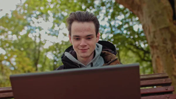 Um rapaz sério, jovem. estudante trabalha em um laptop no parque. Trabalho remoto e conceitos de estudo. Imagem de perto de um jovem programador a alegrar-se com a câmara. O conceito de um adolescente de sucesso no — Fotografia de Stock