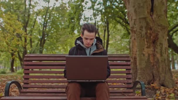 Medium shot of handsome young man working on laptop computer sitting outdoors in park, then looks at the camera and shows OK.Work alone is far from the office,freelancer — Stock Video