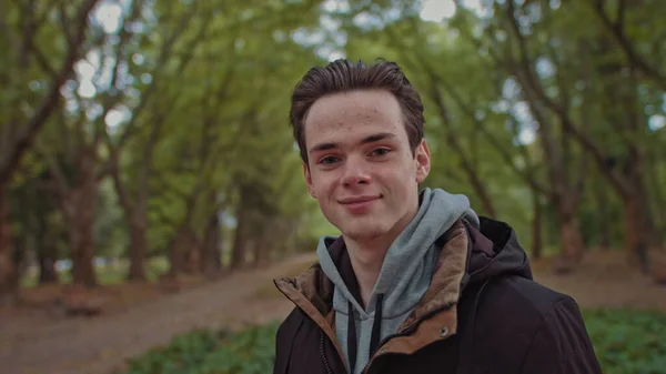 Joven hombre feliz mira a la cámara. Feliz cara masculina sonriente, al aire libre. Primer plano de la cara de los adolescentes. Lentamente acercándose a la cara de los humanos.Un adolescente se para en el parque y disfruta de la vida. Mira derecho. — Foto de Stock