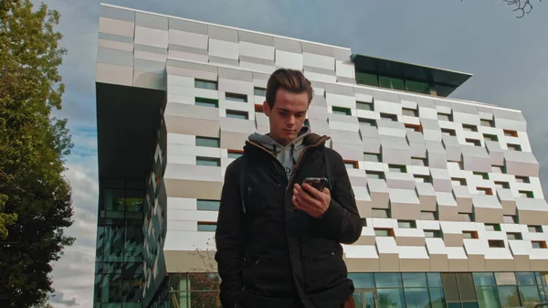 En ung mann, tenåring, som ser på mobiler, snur blikket mot kameraet. Portrett av en tenåringsgutt med bakgrunn fra et forretningssenter, moderne bygning. Mobilt seierskonsept. Munter fyr gjør en – stockfoto