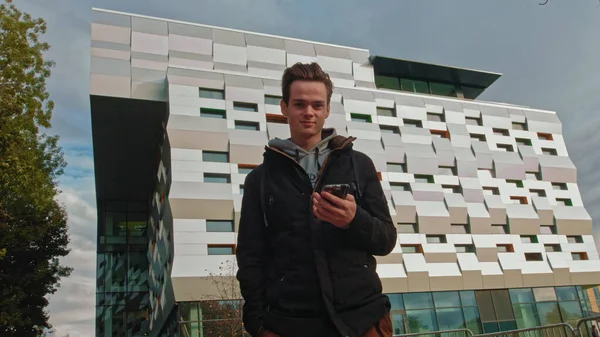 Νεαρός, έφηβος, κοιτάζοντας το κινητό στρέφει το βλέμμα του στην κάμερα. Πορτρέτο ενός έφηβου αγοριού στο παρασκήνιο ενός επιχειρηματικού κέντρου, σύγχρονο κτίριο. Κινητό σχέδιο νίκης. Χαρούμενος τύπος κάνει μια — Φωτογραφία Αρχείου