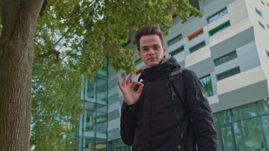 Yakından ateş eden genç bir adamın portresi. Omuzlarında sırt çantası olan şık giyimli bir öğrenci kameraya iyi görünüyor. İş merkezinin arka planında, modern bina