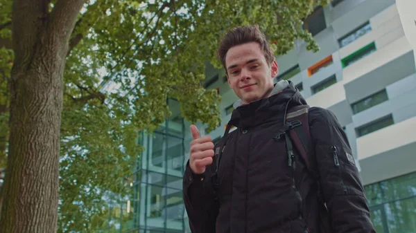 Handsome smiling young guy of 18 years in modern warm clothes on the background of the business center shows like. People sincere emotions, lifestyle concept. Looking approvingly at the camera showing — Stock Photo, Image