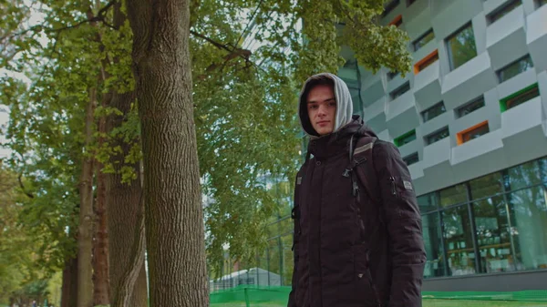 Portrait of a hipster young man putting on a hood while looking at the camera. Close up view of millennial boy in dark hoodie smiling standing on the street. People emotion concept — Stock Photo, Image