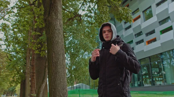 Portrait of a hipster young man putting on a hood while looking at the camera. Close up view of millennial boy in dark hoodie smiling standing on the street. People emotion concept — Stock Photo, Image