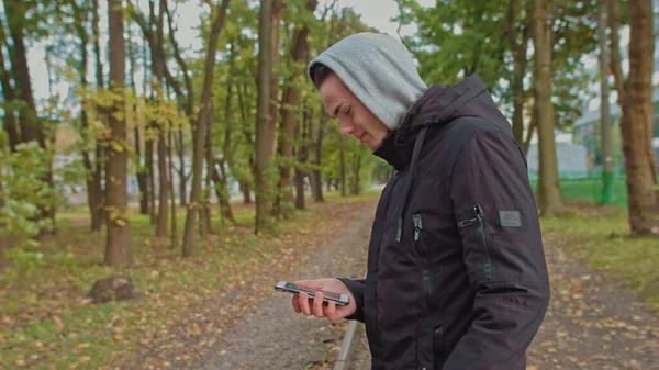 Cortando uma visão de um hipster milenar, abrindo e rolando a tela do telefone. Jovem usando um smartphone enquanto caminha na rua no parque. Um jovem de capuz olha para o telefone — Fotografia de Stock