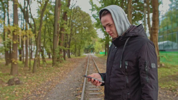 Cortando uma visão de um hipster milenar, abrindo e rolando a tela do telefone. Jovem usando um smartphone enquanto caminha na rua no parque. Um jovem de capuz olha para o telefone — Fotografia de Stock