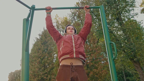 Ung tonårskille gör pull ups övningar på horisontell bar under intensiv träning utomhus. Amerikansk hipster tränar ensam i stadsparken på vårdagen. Aktiv person bär snygg — Stockfoto
