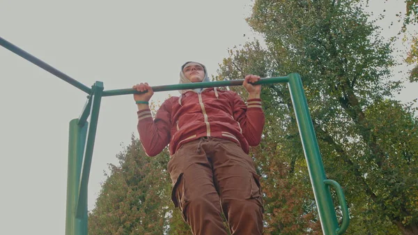Ung tonårskille gör pull ups övningar på horisontell bar under intensiv träning utomhus. Amerikansk hipster tränar ensam i stadsparken på vårdagen. Aktiv person bär snygg — Stockfoto