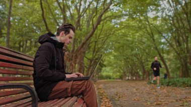 Parkta dizüstü bilgisayarı olan genç bir adam, genç bir adam, parkta dizüstü bilgisayarla çalışan bir öğrenci. Uzak çalışma ve öğrenme kavramı