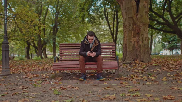 Giovane ragazzo seduto su una panchina del parco con un telefono in mano. giovane albero verde elegantemente vestito al centro della cornice. Il ragazzo usa uno smartphone — Foto Stock