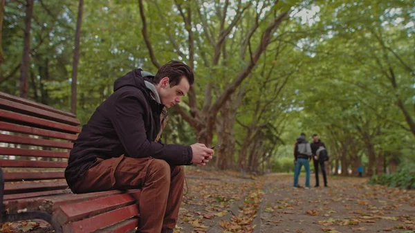 Junger Mann mit Handy in der Hand, der auf einer Bank im Park sitzt. Im Hintergrund eine Gruppe von Freunden, die im Park spazieren gehen. Das Konzept der Einsamkeit und Abhängigkeit von Internet und Smartphones. — Stockfoto