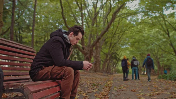 손에 전화기를 들고 공원 벤치에 앉아 있는 청년. 배경에는 친구들이 공원을 걷고 있습니다. 외로움과 인터넷과 스마트폰에 대한 의존의 개념. — 스톡 사진