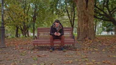 Elinde telefonla parkta bankta oturan genç adam. Yeşil ağaçlı genç adam çerçevenin ortasında şık bir şekilde giyinmiş. Adam akıllı telefon kullanıyor.