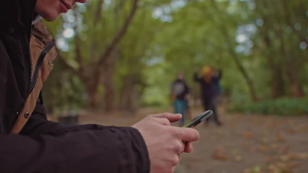 En ung fyr med en telefon i hendene sittende på en benk i parken. I bakgrunnen, en gruppe venner som går i parken. Begrepet ensomhet og avhengighet av Internett og smarttelefoner. – stockfoto