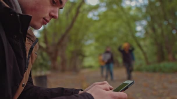 Young guy with a phone in his hands sitting on a bench in the park. In the background, a group of friends walking in the park. The concept of loneliness and dependence on the Internet and smartphones. — Stock Video