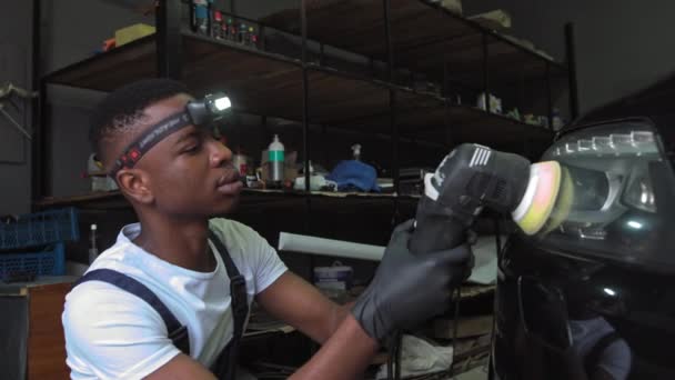 Un joven afroamericano trabaja en el trabajo. Pulido faros del coche. Un hombre utiliza un dispositivo especial para restaurar los faros de un coche negro costoso.Detallando, limpiando la superficie del coche de — Vídeo de stock