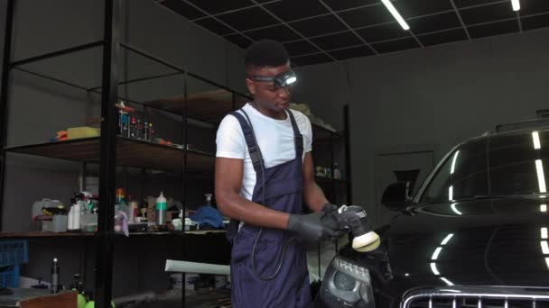 Ein junger Afroamerikaner arbeitet bei der Arbeit. Autoscheinwerfer polieren. Ein Mann benutzt eine spezielle Vorrichtung, um die Scheinwerfer eines teuren schwarzen Autos wiederherzustellen. — Stockvideo