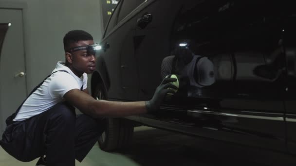 Um trabalhador de serviço afro-americano aplica uma camada protetora de leocerâmicos.Polimento do farol, processamento de luzes do carro. Um trabalhador do serviço de carro polir o farol de um carro de passageiros. Profissional — Vídeo de Stock