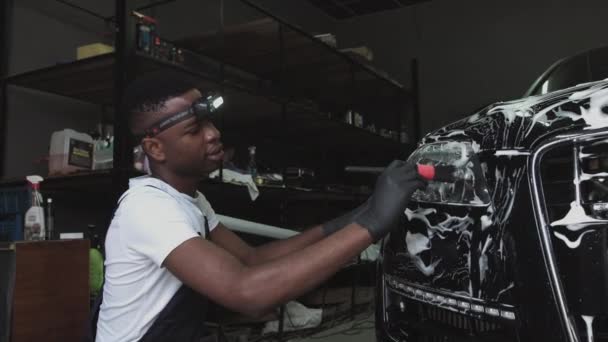 Giovane studente al lavoro. Un afro-americano lavora in un autolavaggio, guadagnando soldi dopo l'allenamento. Un uomo in abiti da lavoro speciali pulisce l'auto con una spazzola. La macchina è in schiuma. — Video Stock