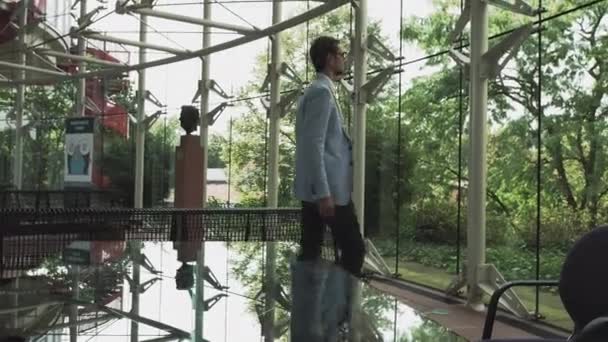 Um jovem bonitão em um terno de arquiteto ou advogado anda em torno de um escritório moderno. Reflexão em uma mesa de vidro. — Vídeo de Stock