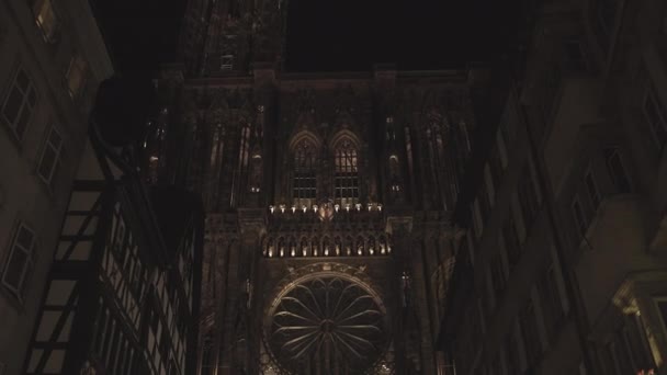 Estrasburgo, Francia - 8 de mayo de 2020: Cámara push-in hacia adelante inclinada desde la entrada a la torre de la fachada frontal iluminada de la catedral de Notre Dame en el casco antiguo de Petite France por la noche entre edificios — Vídeos de Stock